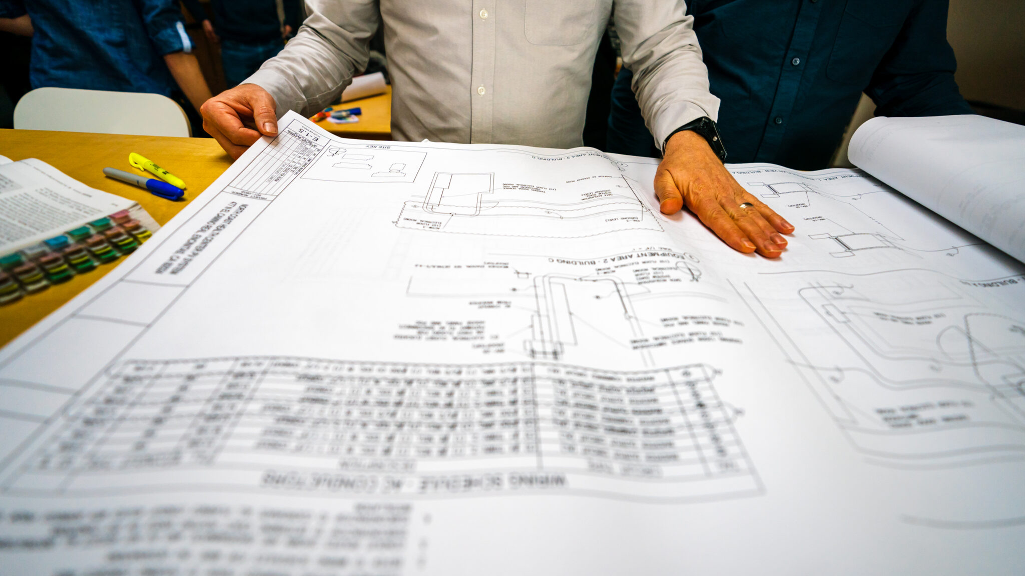 Hands spread across large electrical drawings on a table, reviewing a wiring schedule and conduit plan