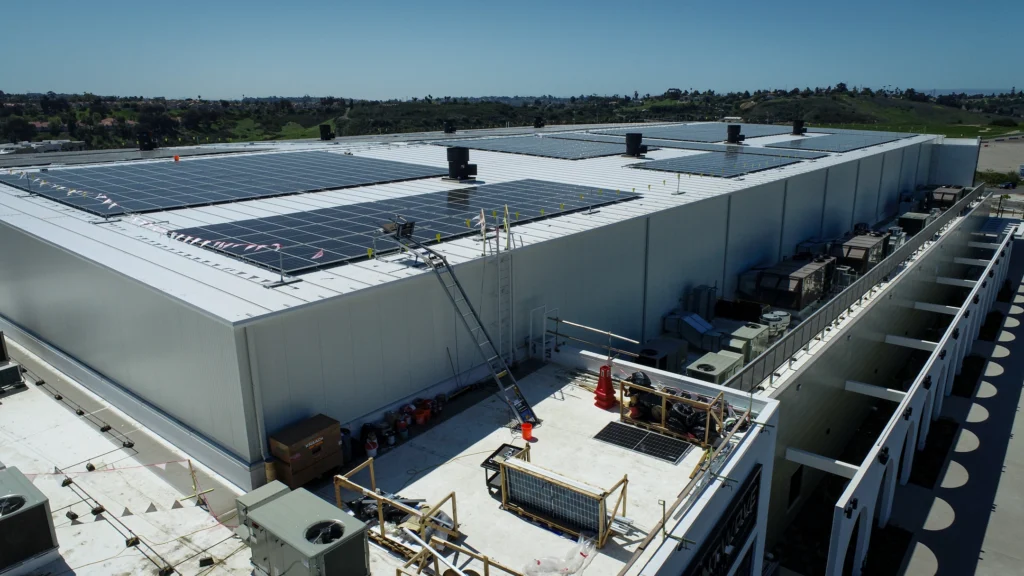 This image shows the rooftop of a large building with solar panels installed. The panels are arranged in rows, covering much of the roof, indicating a significant investment in renewable energy. The area also appears to be a construction or maintenance zone, with tools, a ladder, and equipment scattered around.