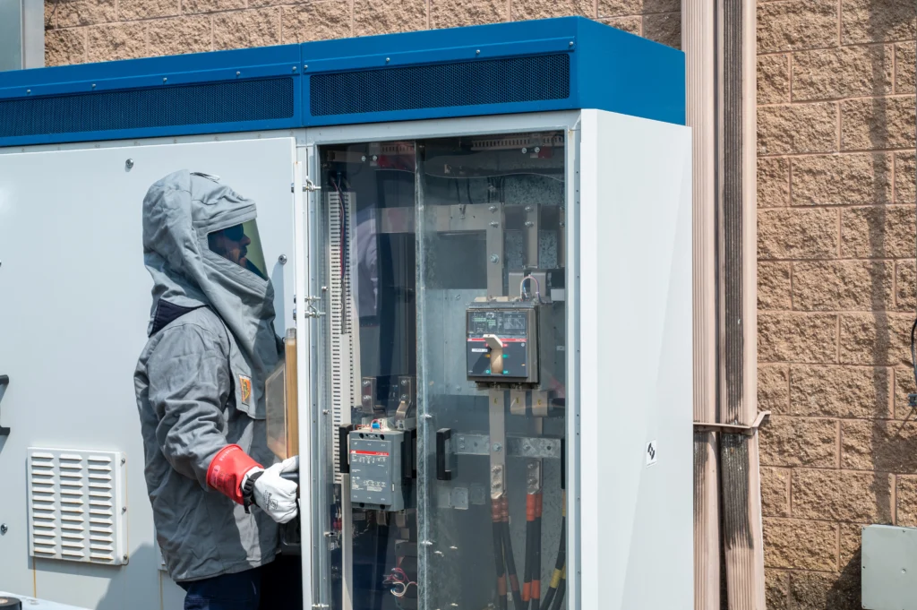 Technician in arc flash PPE inspecting electrical panel outdoors